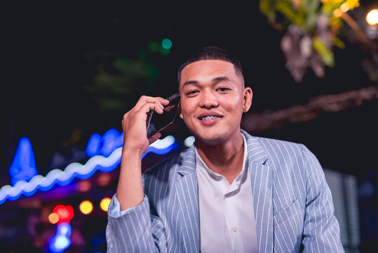 Portrait Of A Handsome And Dapper Young Man Hanging Out At An Outdoor Bar. Holding A Phone Listening To Someone. Nightlife Scene At A Popular Spot.