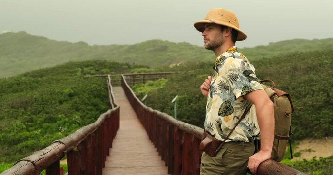 Traveling along mountains and rain forest, freedom and active lifestyle concept. back view male traveler walks along a wooden bridge in the jungle near the ocean wearing a hat and a backpack