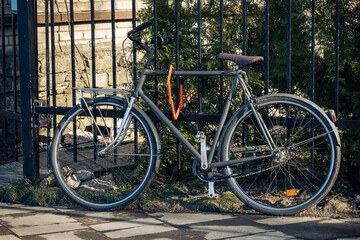 Black retro bicycle parked against metal fence with house wall and garden in the background....