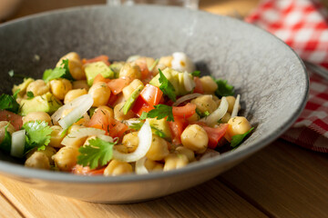 45º view of a chickpeas salad with avocado, tomato, coriander and onion, served on a grey dish with a red chekered cloth on a wooden table. Horizontal image