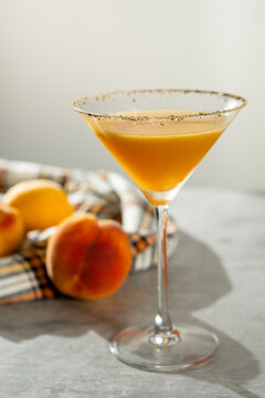 Peach margarita served in a martini glass with pepper. It sits on a grey surface with a checkered cloth and peaches, with a white background and hard light. Vertical image