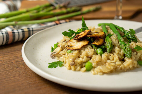 45º view of a mushroom and asparagus risotto served on a white dish with a fork, a glass of white wine and a grater and asparagus on a cutting board, surrounded by a checkered cloth. Horizontal image