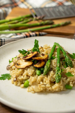 45º view of a mushroom and asparagus risotto served on a white dish with a fork, a glass of white wine and a grater and asparagus on a cutting board, surrounded by a checkered cloth. Vertical image