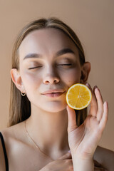 portrait of pleased woman with closed eyes holding half of juicy lemon isolated on beige.