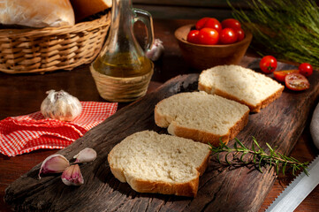 fresh and homemade whole and sliced bread with oil can and cherry tomatoes wicker pan and rosemary on wooden table with light from a window