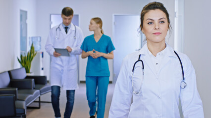 In a modern hospital corridor in front of the camera the mature woman doctor walking through the camera background her colleagues main doctor man and young nurse have a conversation they discussing