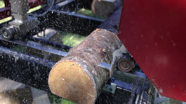 Short clip of an industrial band saw at work outdoors, removing the outer layer and squaring off freshly chopped down trees to make wooden planks.