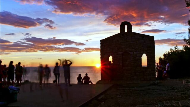 Sunset timelapse 4K with black silhouettes of unrecognizable people and lovers embraced. Pula Istria Croatia Sacred rock Sveta Stijena a wall of an church on Verudela Peninsula. Romantic love concept