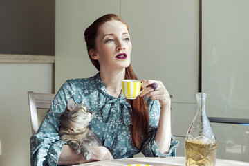beautiful elegant woman with a cat sits in the kitchen  and holds a teacup with wine