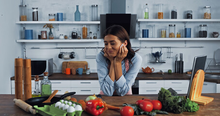 brunette woman smiling near fresh vegetables and cooking utensils on kitchen worktop.