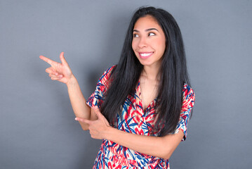 Optimistic Young latin woman wearing colorful dress over grey backgrogund points with both hands...