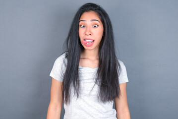 Obraz premium Funny young beautiful brunette woman wearing white t-shirt over grey background makes grimace and crosses eyes plays fool has fun alone sticks out tongue.