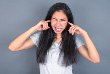 Happy young beautiful brunette woman wearing white t-shirt over grey background ignores loud music and plugs ears with fingers asks to turn off sound