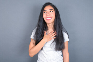 Fototapeta premium Joyful young beautiful brunette woman wearing white t-shirt over grey background expresses positive emotions recalls something funny keeps hand on chest and giggles happily.