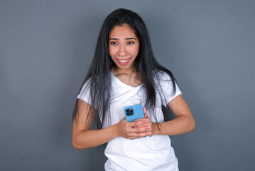 Smiling young beautiful brunette woman wearing white t-shirt over grey background friendly and happily holding mobile phone taking selfie in mirror.