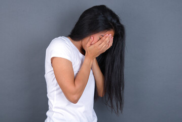 Sad young beautiful brunette woman wearing white t-shirt over grey background covering face with...