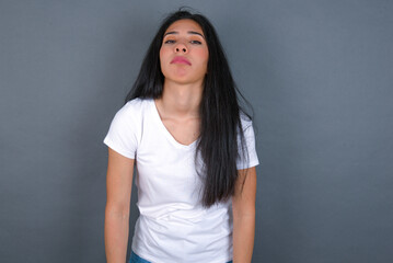 Gloomy, bored young beautiful brunette woman wearing white t-shirt over grey background frowns face looking up, being upset with so much talking hands down, feels tired and wants to leave.