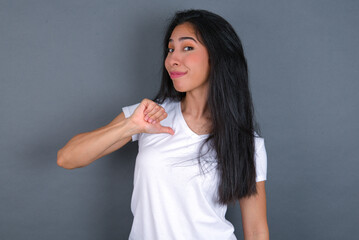 Closeup of cheerful young beautiful brunette woman wearing white t-shirt over grey background looks joyful, satisfied and confident, points at himself with thumb.