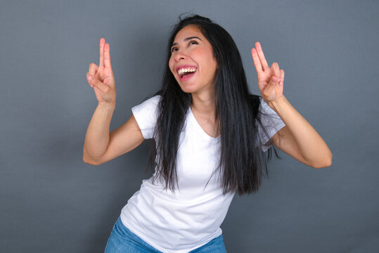 Isolated shot of cheerful young beautiful brunette woman wearing white t-shirt over grey background makes peace or victory sign with both hands, feels cool.