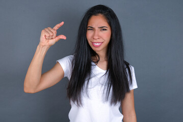 young beautiful brunette woman wearing white t-shirt over grey background smiling and gesturing with hand small size, measure symbol.