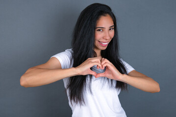 Fototapeta premium young beautiful brunette woman wearing white t-shirt over grey background smiling in love showing heart symbol and shape with hands. Romantic concept.