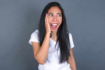 young beautiful brunette woman wearing white t-shirt over grey background excited looking to the...