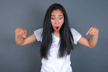 Amazed young beautiful brunette woman wearing white t-shirt over grey background points down with...
