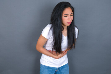 young beautiful brunette woman wearing white t-shirt over grey background suffering from strong stomachache.