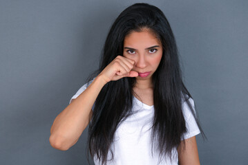 Unhappy young beautiful brunette woman wearing white t-shirt over grey background crying while posing at camera whipping tears with hand.