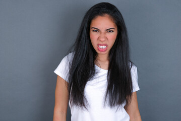 Mad crazy young beautiful brunette woman wearing white t-shirt over grey background clenches teeth...