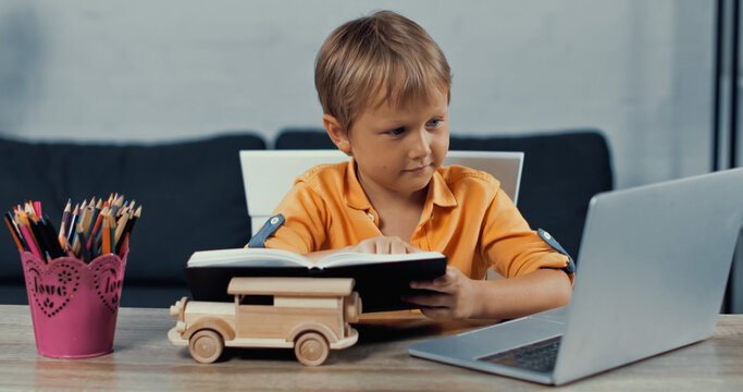 boy holding notebook and looking at laptop near wooden toy car on desk.