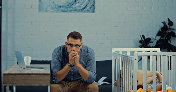 worried father in eyeglasses sitting near baby crib with infant son and desk with gadgets.