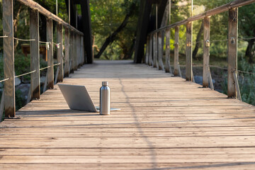 Isolated front view of laptop and metl bottle of water on a wooden flooring with nature in the background and copy space