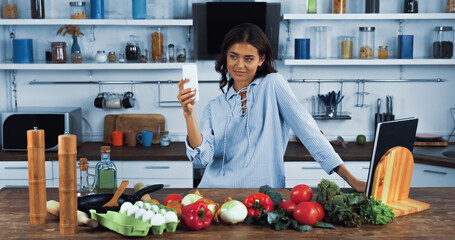 happy woman taking selfie on smartphone near raw ingredients on kitchen table.