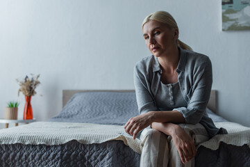 upset blonde woman with menopause sitting on bed at home.