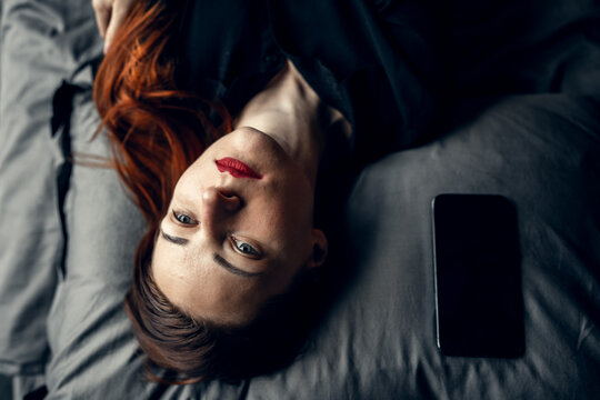 Sad young redhead woman lying on pillow with mobile phone and waiting for call or message.