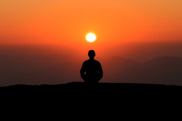 Naklejka na ściany i meble Silhouette of young asian man practices yoga and meditates alone on top of the mountain with beautiful view orange sky and sunrise at morning.