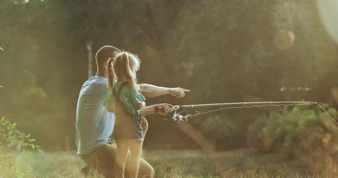 Dad throws fishing rod into river to catch fish and blonde daughter helps. Father with little girl has rest together on blurred background