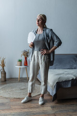 full length of blonde woman with menopause suffering from heat and holding fan in bedroom.