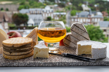 Cow cheeses of Normandy - camembert, livarot, neufchatel, pont l'eveque and glass of apple cider drink with houses of Etretat village on background, Normandy, France