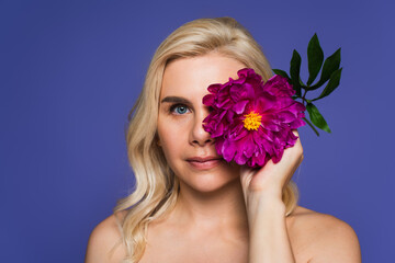 blonde woman with bare shoulders covering eye with blooming flower and looking at camera isolated on purple.