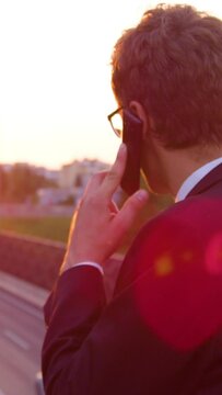 SLOW MOTION, LENS FLARE, CLOSE UP: Successful young businessman making an important phone call on a sunny evening. Unrecognizable agent in a suit calling his client while observing the busy highway.