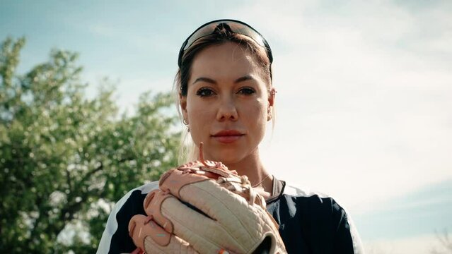 A dramatic close up of a  female softball athlete posing with her baseball glove on a practice field.  She stares intently into the camera producing an epic atmosphere. Shot in glorious slow motion.