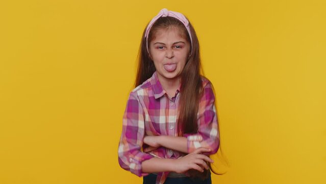 Cheerful funny young preteen child girl kid showing tongue making faces at camera, fooling around, joking, aping with silly face, teasing. Little toddler children isolated on studio yellow background