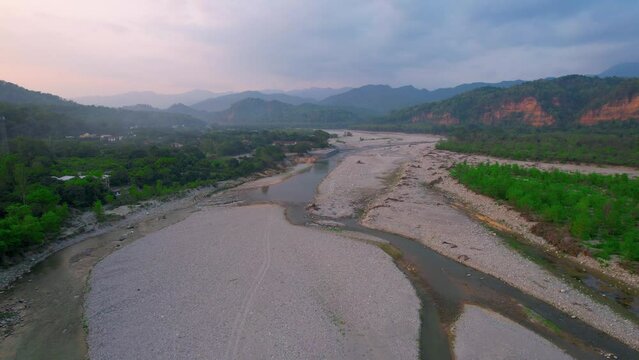 aerial drone shot flying towards large fog covered mountains in the distance over a dried river bed with green trees on both sides at jim corbett park in india a popular tourist destination