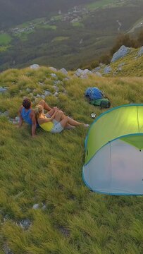 AERIAL: Flying above the carefree tourist couple observing the evening landscape after a mountain biking journey in the green Alps. Active man and woman cuddling by their tent in the scenic mountains.