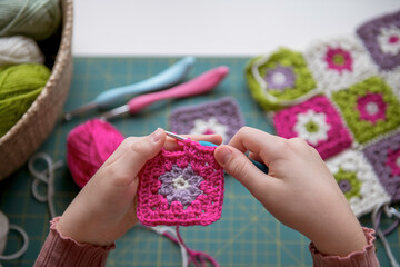 Girl's hands crochetting with colorful yarn granny squares blanket. Hobby crafting and handmade. Overhead creative workspace with cutting mat, scissors and crochet hooks