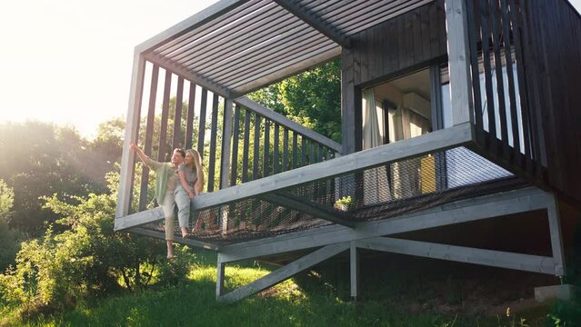 Young couple sitting and cuddling in hammock terrace in their new home in tiny house in woods, sustainable living concept.