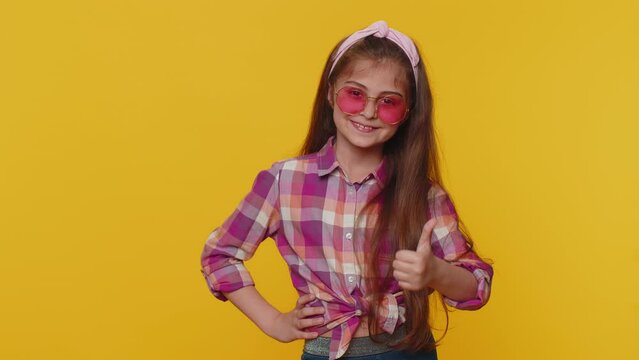 Portrait of seductive preteen child girl kid wearing pink sunglasses, charming smile, showing thumbs up like, positive feedback. Little stylish toddler children isolated on studio yellow background