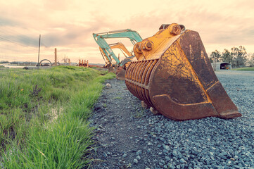 View of a parking lot of construction equipment on cloudy background of a sunset summer day. Pink gray clouds. Old rusty excavator bucket close-up.